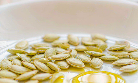 Macro of glossy green pumpkin seeds scattered across white ceramic beside a shallow pool of golden oil