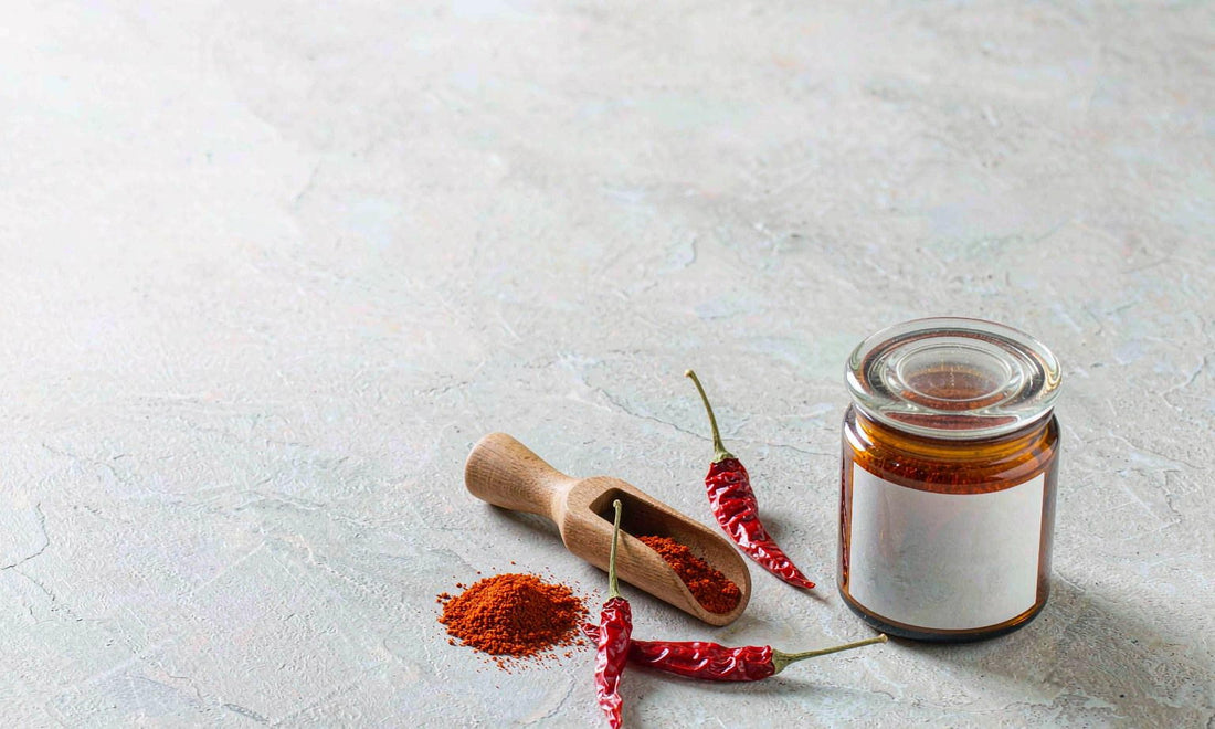 Flat lay of ground cayenne powder, three dried red chillies and a blank amber jar on pale grey stone