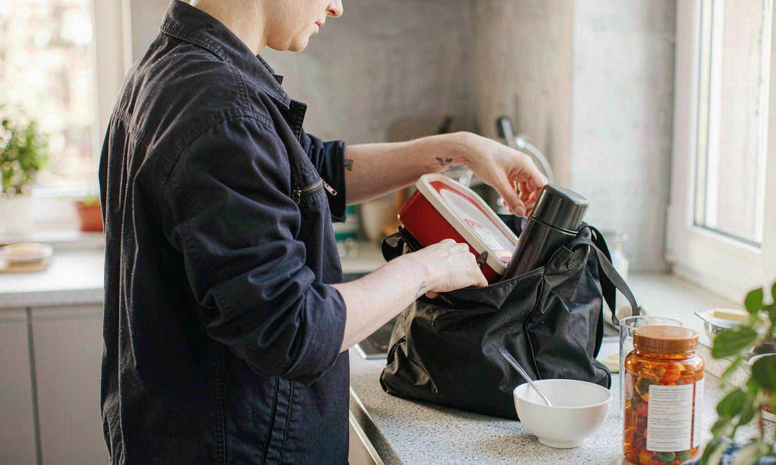 A shift worker seen from behind packing a lunchbox and thermos into a work bag on a pale dawn kitchen counter