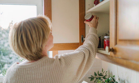 A person seen from behind in a cream sweater placing a small jar into a bright kitchen cupboard