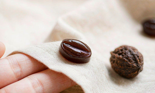 Macro close-up of a dark botanical gummy resting on pale linen with fingertips and a walnut shell nearby
