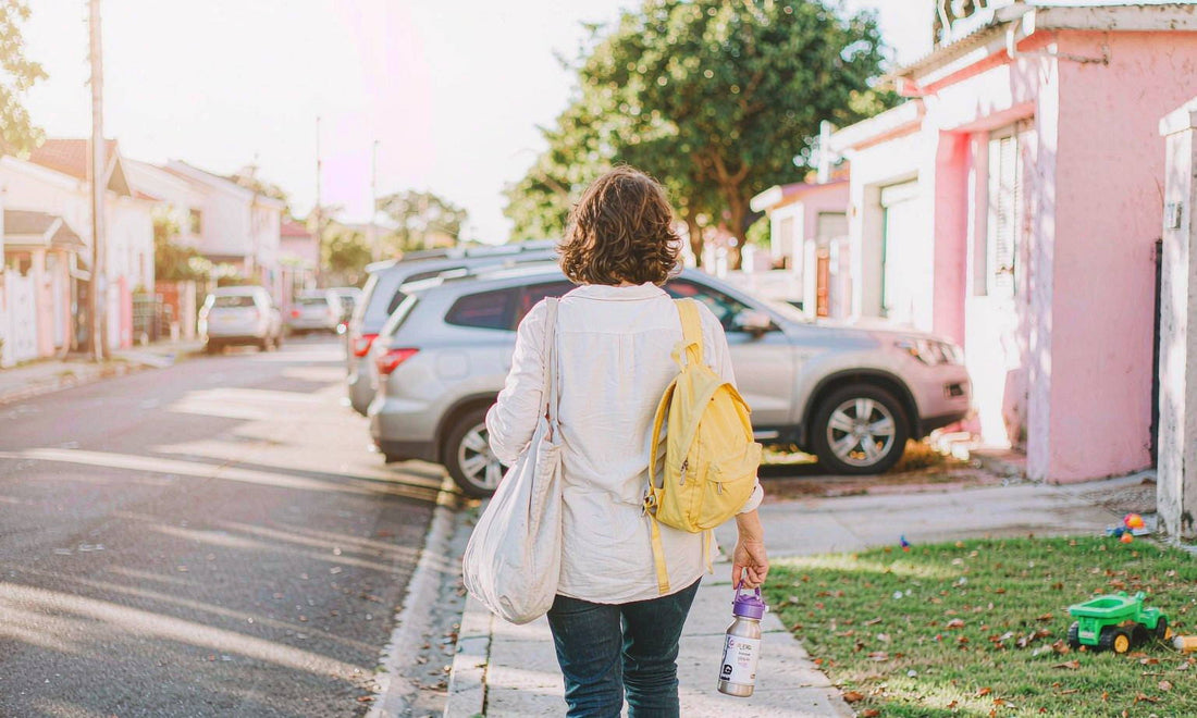 A woman seen from behind on a sunlit sidewalk with a canvas tote, a child's backpack and a water bottle
