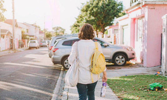 A woman seen from behind on a sunlit sidewalk with a canvas tote, a child's backpack and a water bottle