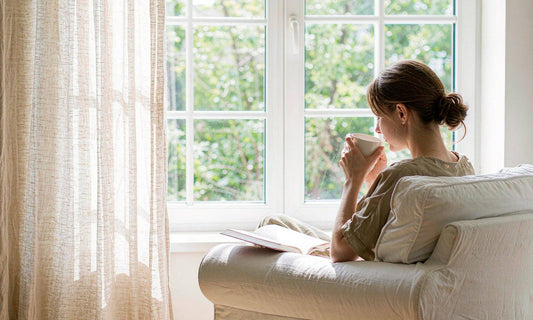 A woman seen from behind resting in a pale armchair by a bright window with a mug of tea and an open book