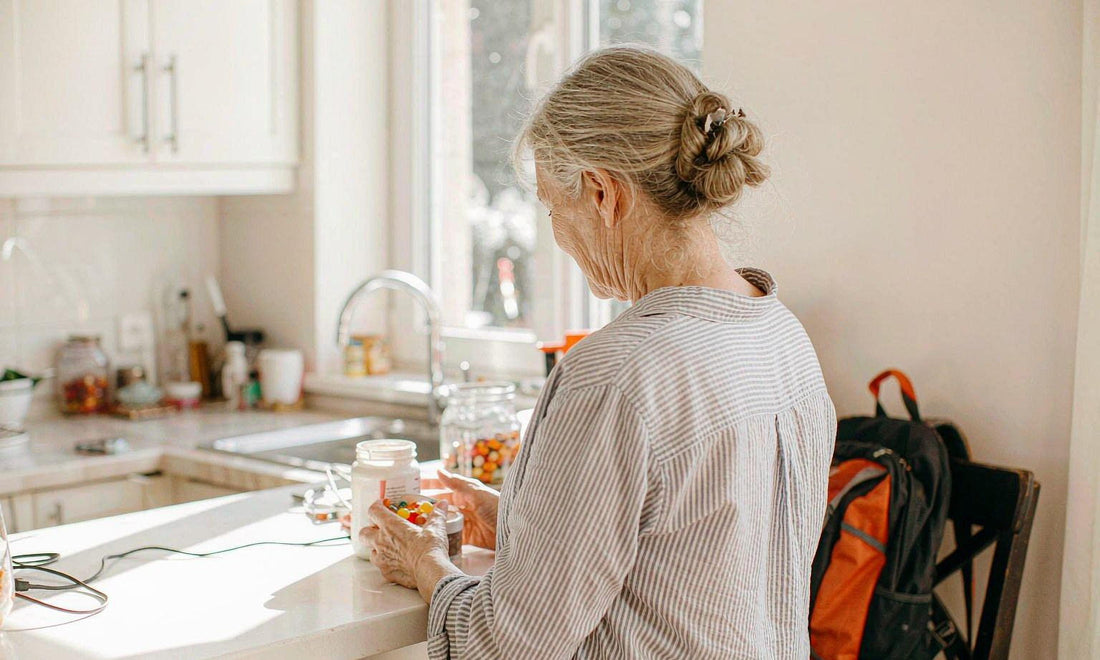 Older woman seen from behind holding a white gummy jar at a sunlit kitchen counter with a school backpack