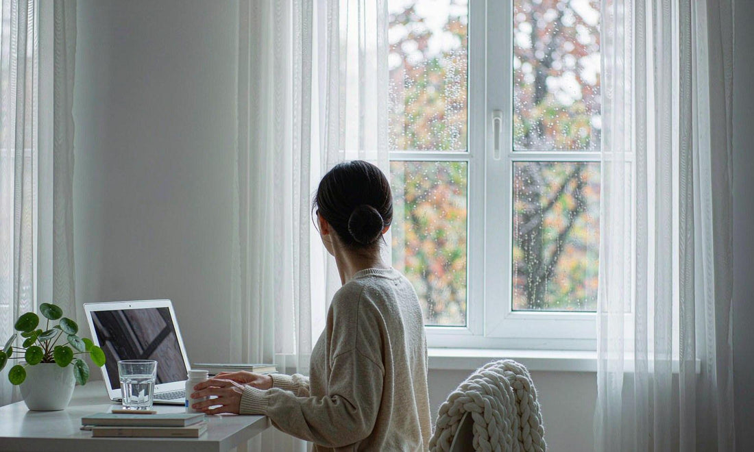 A person seen from behind at a bright home office desk with a laptop, a glass of water and rainy windows
