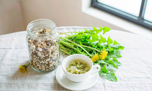 A glass jar of dried dandelion root beside a cup of herbal tea, fresh greens and yellow flowers on linen