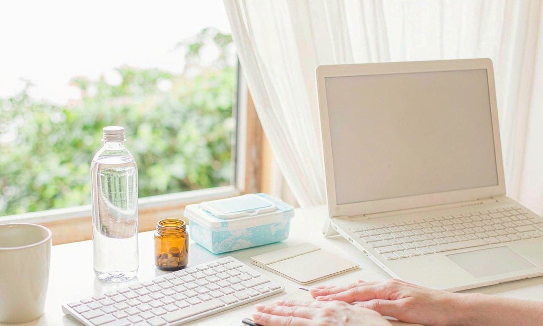 A tidy home office desk in morning light with a laptop, water bottle, amber jar and a pack of wipes