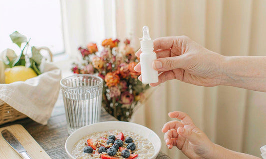 An adult hand holding a dropper bottle above a breakfast table with oatmeal and a glass of water