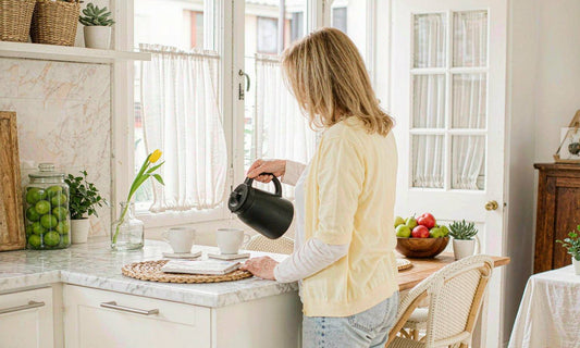 A woman in her fifties seen from behind pouring coffee at a sunlit white marble kitchen counter