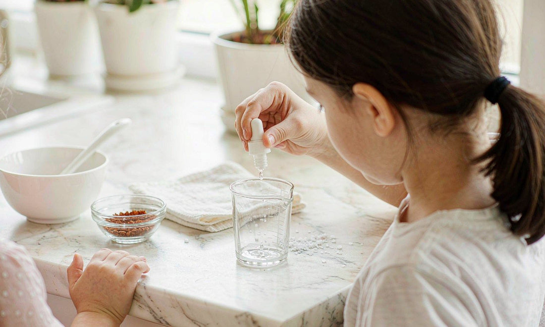 A parent seen from behind holding a dropper bottle over a measuring cup on a bright kitchen counter
