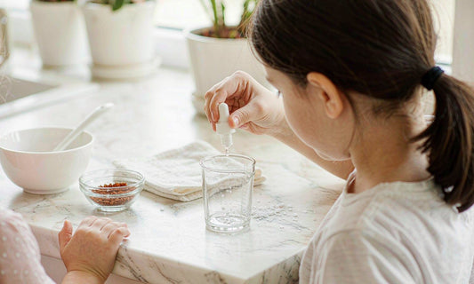 A parent seen from behind holding a dropper bottle over a measuring cup on a bright kitchen counter