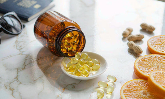 Golden softgels spilling from an amber jar into a small dish with orange slices and peanuts on marble