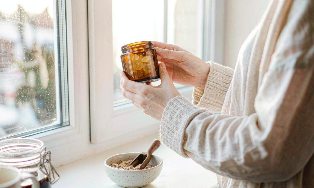 Hands holding an unlabelled amber jar of gummies up to a sunlit window above a bowl of powder