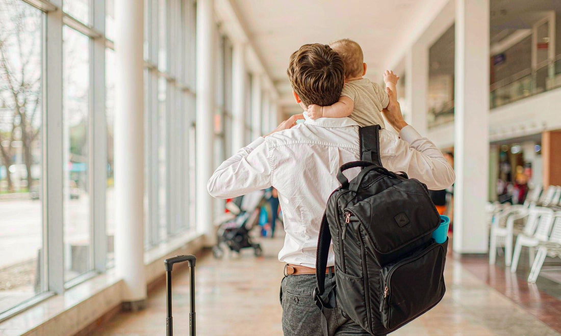 Back view of a parent carrying a baby through a sunlit airport terminal with suitcase and diaper backpack