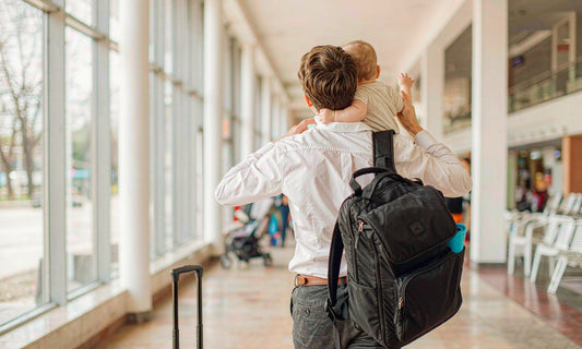 Back view of a parent carrying a baby through a sunlit airport terminal with suitcase and diaper backpack