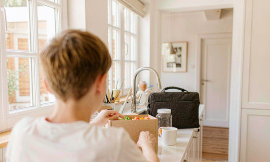 Parent packing school lunchboxes on a sunlit kitchen counter with a jar of gummies beside a laptop bag