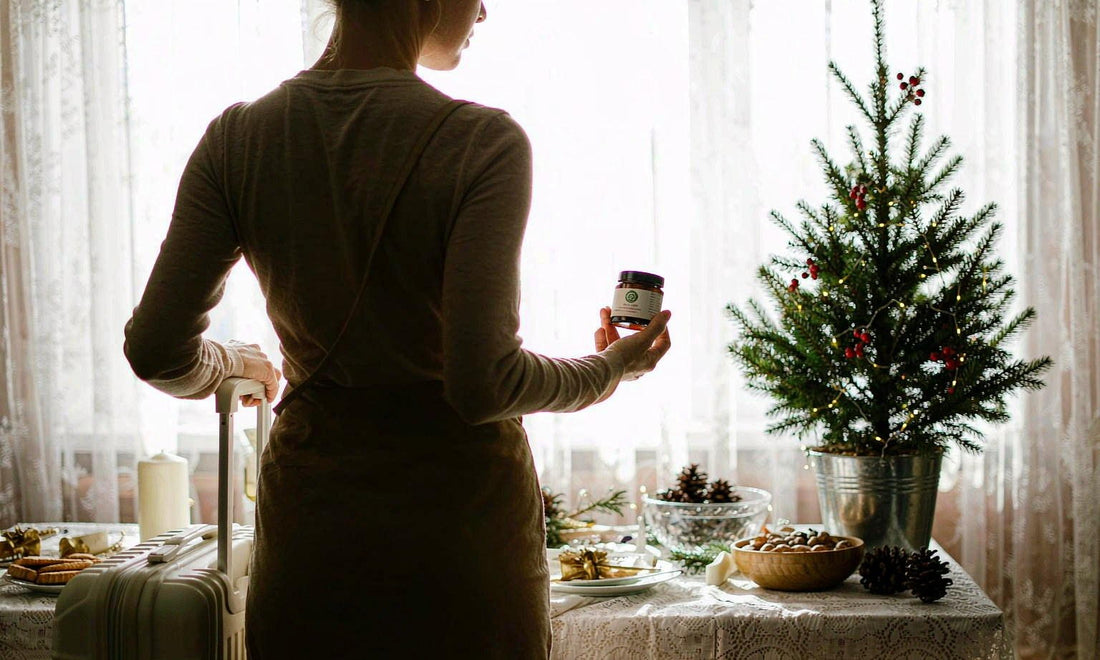 A traveler seen from behind holding a small amber jar beside a decorated holiday tree with warm lights