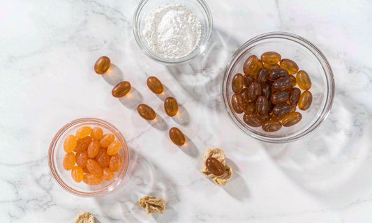 Top-down flat lay of amber gummies, pectin powder and dried fruit in glass bowls on white marble