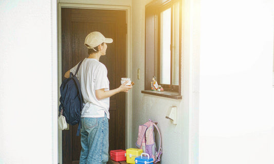 A parent seen from behind at a bright doorway with a backpack and lunchboxes waiting by the door