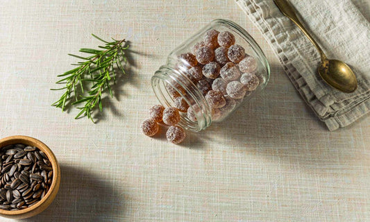 Overhead flat-lay of a glass jar of amber whole-food gummies with rosemary, seeds and linen on pale cloth