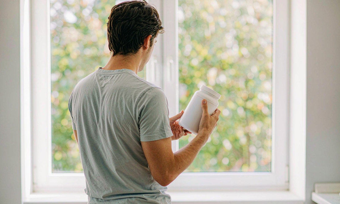 A man seen from behind holding a white supplement jar at a sunny kitchen window in the morning