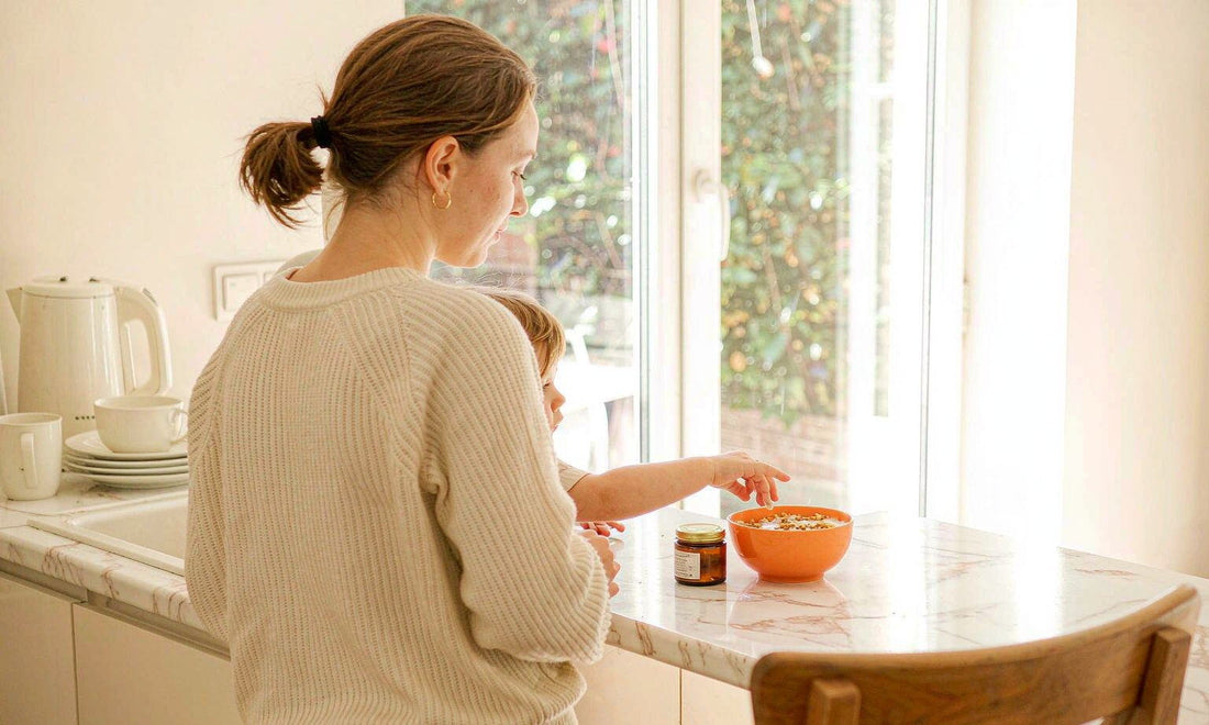 A woman placing a small amber tin beside a child's breakfast bowl on a sunlit kitchen counter