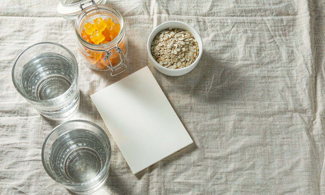 Overhead flat lay of a gummy jar on linen beside a bowl of oats, a glass of water and a small notebook