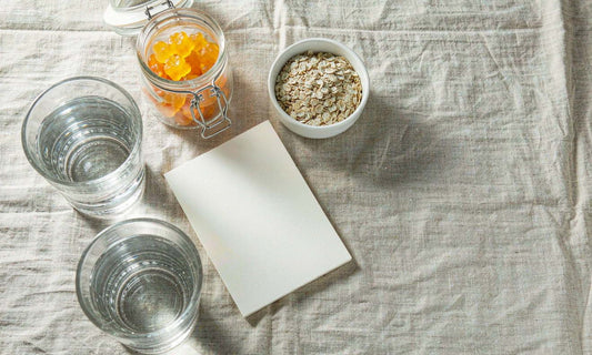 Overhead flat lay of a gummy jar on linen beside a bowl of oats, a glass of water and a small notebook