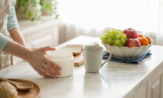 A hand setting a white supplement jar on a marble kitchen counter beside a coffee mug and fruit bowl
