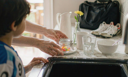 An adult's hands setting a small gummy jar on a breakfast counter beside a child's soccer gear and cleats