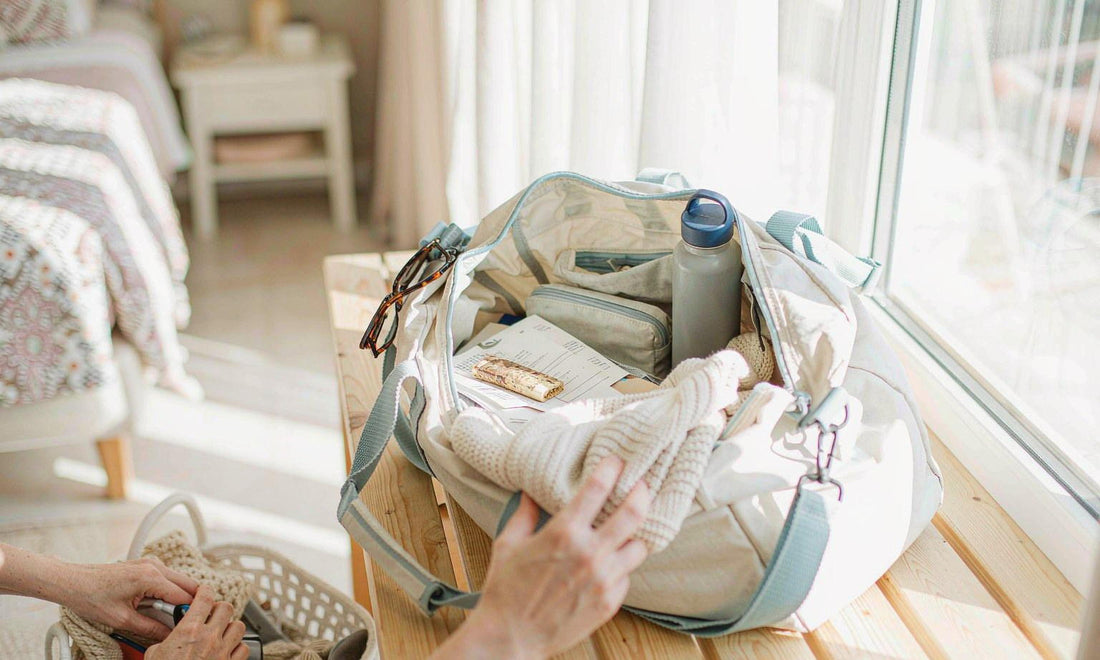 An open canvas travel bag packed with a water bottle, pouch and folded list on a wooden bench