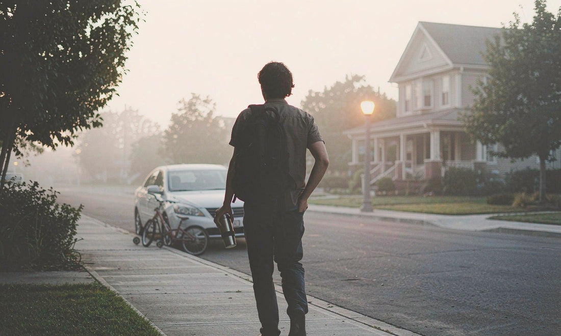 Commuter from behind with backpack and coffee cup walking toward golden sunrise light on a quiet street